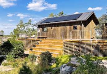 Chalet en bois avec panneaux solaires et grande terrasse à VÄDDÖ, Suède, entouré de nature et ciel bleu.
