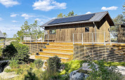 Wooden cabin with solar panels and spacious deck in VÄDDÖ, Sweden, surrounded by trees and blue sky.