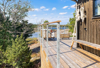 Terrasse en bois avec vue sur l’eau et la forêt à une cabane à VÄDDÖ, Suède sous un ciel ensoleillé.