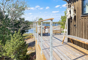 Terrasse en bois avec vue sur l’eau et la forêt à une cabane à VÄDDÖ, Suède sous un ciel ensoleillé.