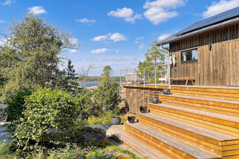 Terrasse en bois et cabane moderne avec vue sur la forêt et l’eau sous ciel bleu à Väddö, Suède.