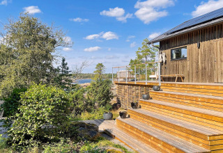 Terrasse en bois et cabane moderne avec vue sur la forêt et l’eau sous ciel bleu à Väddö, Suède.
