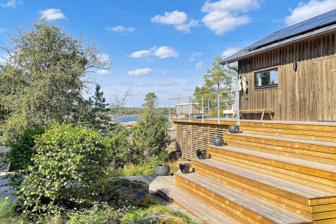 Terrasse en bois et cabane moderne avec vue sur la forêt et l’eau sous ciel bleu à Väddö, Suède.