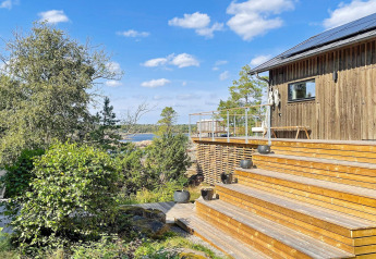 Terraza de madera y cabaña moderna con vistas al bosque y al agua bajo un cielo azul en Väddö, Suecia.