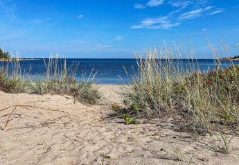 View from a cabin at VÄDDÖ, Sweden, showing sandy beach, dune grass, and calm blue sea under clear sky.
