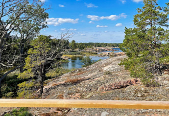 Vista da una baita a VÄDDÖ, Svezia, con rocce, alberi e acqua sotto un cielo azzurro.