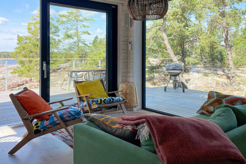 Wohnzimmer eines Ferienhauses in VÄDDÖ, Schweden, mit bunten Kissen, großen Fenstern und Waldblick.