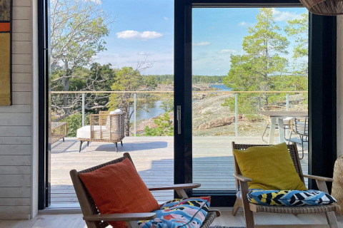 Blick aus dem Wohnzimmer eines modernen Ferienhauses in VÄDDÖ, Schweden, mit Terrasse, Stühlen und Seeblick.