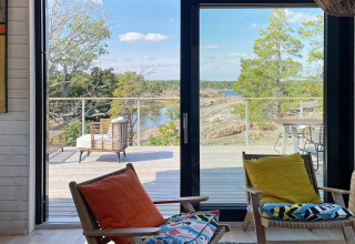 Zicht vanuit een vakantiehuis in VÄDDÖ, Zweden, met terras, stoelen, bomen en een waterpartij op de achtergrond.