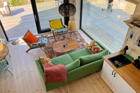 Salon lumineux dans une cabane suédoise avec canapé vert, coussins colorés et grandes baies vitrées sur la terrasse.
