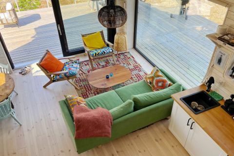Helles Wohnzimmer in einer schwedischen Hütte mit grüner Couch, farbenfrohen Kissen und Holzterrasse.
