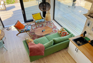 Sala luminosa en una cabaña sueca con sofá verde, cojines coloridos y grandes puertas de vidrio a la terraza.