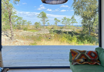 Udsigt fra en moderne hytte i VÄDDÖ, Sverige, med træterrasse, skov og hav i baggrunden.