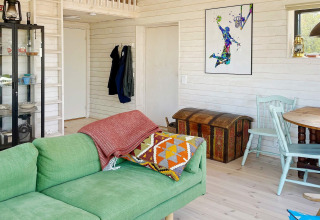 Soggiorno di una baita con divano verde, arte colorata, baule antico e pareti in legno chiaro a VÄDDÖ, Svezia.
