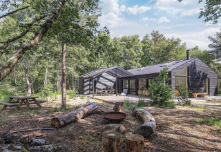 Moderno lodge nero tra i boschi a Læsø, Danimarca, con area fuoco esterna e sedute in tronchi.