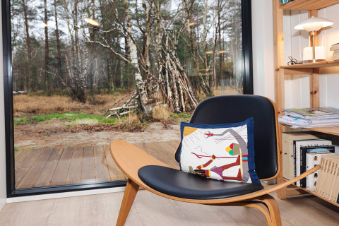 Moderner Stuhl mit bunter Kissen vor großen Fenstern mit Blick auf den Wald im 5-Sterne-Haus auf Læsø.