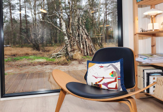 Moderner Stuhl mit bunter Kissen vor großen Fenstern mit Blick auf den Wald im 5-Sterne-Haus auf Læsø.