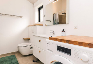 Moderne badeværelse med vaskemaskine, træbordplade, væghængt toilet og stort spejl i Læsø feriehus.