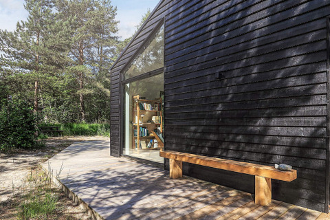 Façade noire moderne d’un lodge avec grande baie vitrée et banc en bois à Læsø, Danemark.