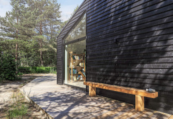 Façade noire moderne d’un lodge avec grande baie vitrée et banc en bois à Læsø, Danemark.