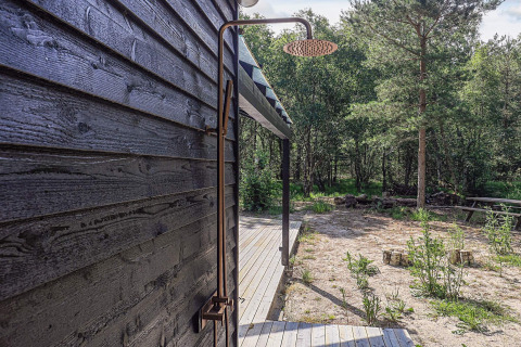 Douche extérieure fixée à un mur de bois noir d’une maison de vacances à Læsø, Danemark, entourée d’arbres.
