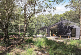 Lodge moderne en bois noir à Læsø, Danemark, entouré d'arbres, avec de grandes fenêtres et un banc extérieur.