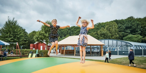 Two children jumping on a large bouncy mat at Horsens City Camp in Denmark with greenery and park buildings.