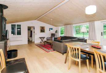 Åben stue med pejs, spisebord og sofa i et lyst sommerhus med store vinduer ud til grøn natur i Holbæk.