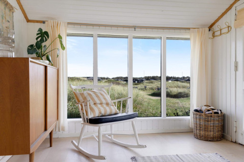 Coin lumineux d'une maison de plage avec fauteuil à bascule, grande fenêtre et vue sur les dunes à Løkken.