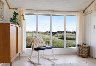 Helles Wohnzimmer in einem Strandhaus mit Schaukelstuhl, großem Fenster und Blick auf die Dünen in Dänemark.