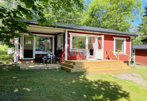 Hyggelig rød hytte ved søen i Sollentuna med terrasse, udendørs siddeområde og omgivet af grønne træer.