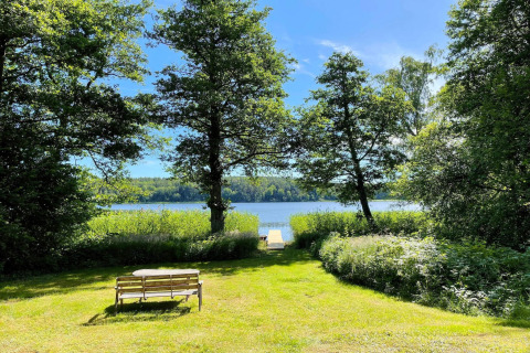 Vue du Lakeside Cabin Retreat à Sollentuna, Suède, avec banc, arbres verts et lac sous un ciel bleu.