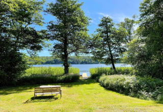 Vue du Lakeside Cabin Retreat à Sollentuna, Suède, avec banc, arbres verts et lac sous un ciel bleu.