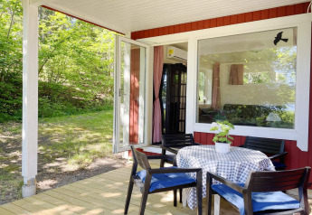 Terrasse en bois avec table et chaises devant un chalet rouge au bord du lac à Sollentuna, Suède, entouré d’arbres.