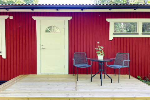 Cabaña roja junto al lago con puerta blanca, terraza pequeña, dos sillas y planta en Sollentuna, Suecia.