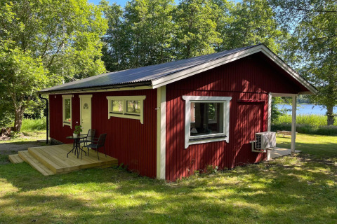 Cabaña roja junto al lago con terraza, rodeada de árboles verdes en Sollentuna, Suecia, en un día soleado.