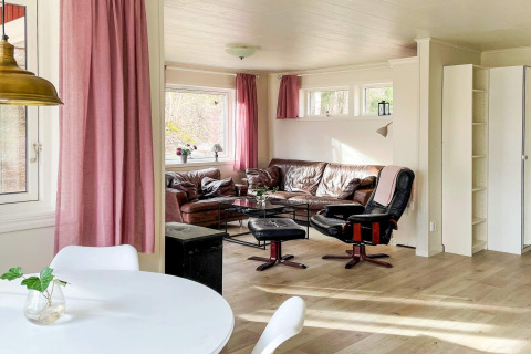 Lyst og hyggeligt opholdsrum i en svensk bjælkehytte med lyserøde gardiner, lædersofa og store vinduer.