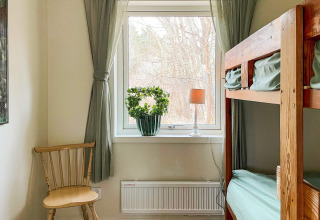 Helles Zimmer mit Etagenbett, grünen Vorhängen, Holzstuhl und Pflanze am Fenster in einer Hütte.