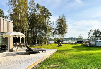 Sonnige Holzterrasse mit Sonnenschirm am Ferienhaus in Hestra, Schweden, mit weitem Blick auf Wiese und See.