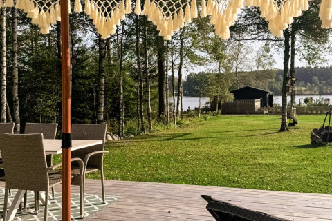 Terrasse med havemøbler, udsigt over græsplæne, træer og sø ved 4-stjernet feriehus i Hestra, Sverige.