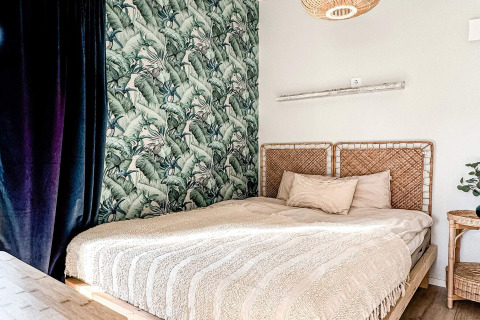 Dormitorio en una cabaña con cabecero de mimbre, papel tropical y luz suave en Hestra, Suecia.