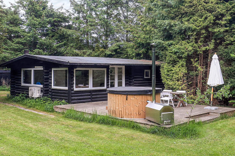 Zwarte houten lodge met hottub, terrasmeubilair en parasol, omringd door groen in Idestrup, Denemarken.