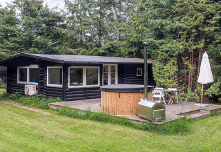 Sort træhytte med stor terrasse, udendørs bad, havebord og parasol, omgivet af grøn have og skov i Idestrup.