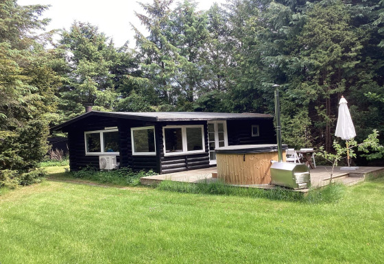Chalet noir en bois pour six personnes à Idestrup, entouré d’arbres, terrasse, bain nordique et parasol.