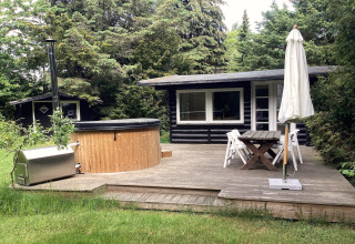 Træterrasse med spabad, havebord og parasol foran en sort træhytte i grønne omgivelser i Idestrup, Danmark.