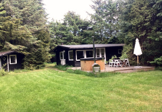 Sort træferiehus med terrasse, spisebord og grøn have i Idestrup, Danmark, omgivet af store træer.