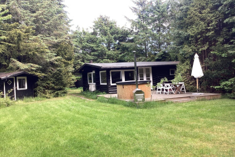 Black wooden holiday lodge with deck, dining area, and grassy yard in Idestrup, Denmark, surrounded by trees.
