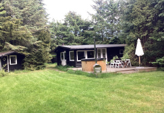 Black wooden holiday lodge with deck, dining area, and grassy yard in Idestrup, Denmark, surrounded by trees.