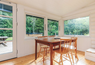 Lyst spisestueområde i et sommerhus i Idestrup, Danmark, med træmøbler og udsigt til grøn natur.