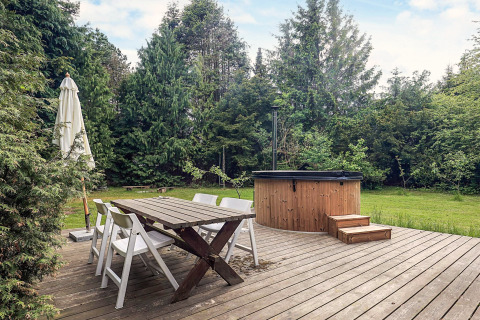 Træterrasse med spisebord, stole, parasol og udendørs badekar omgivet af grønne træer i Idestrup.